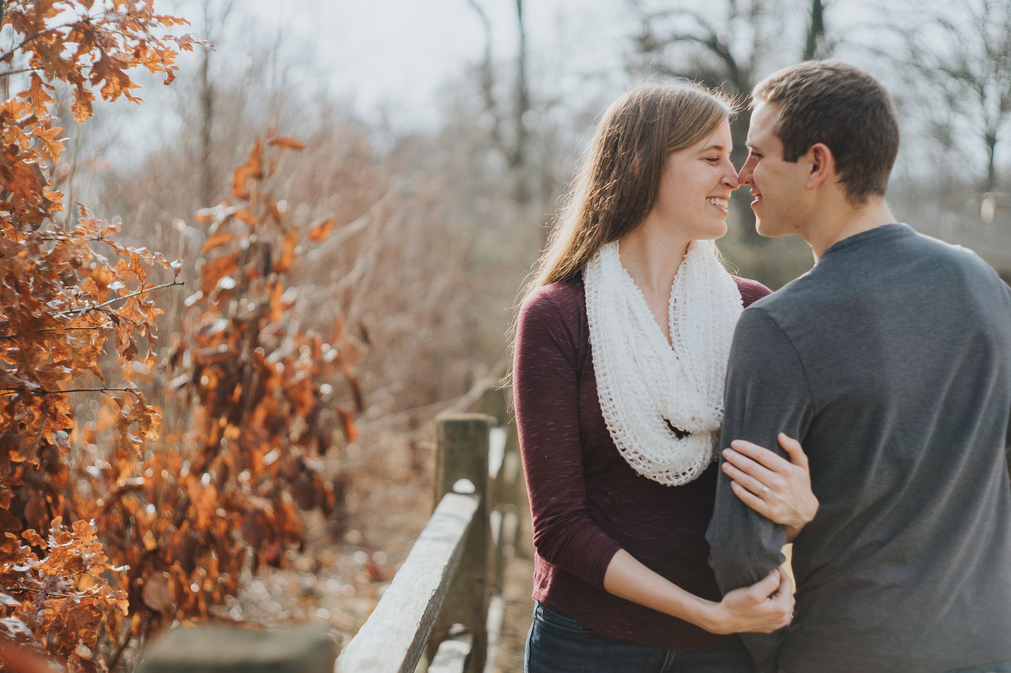 A couple stand near a red tree. They're smiling and touching noses during their engagement session at Blackburn Park.