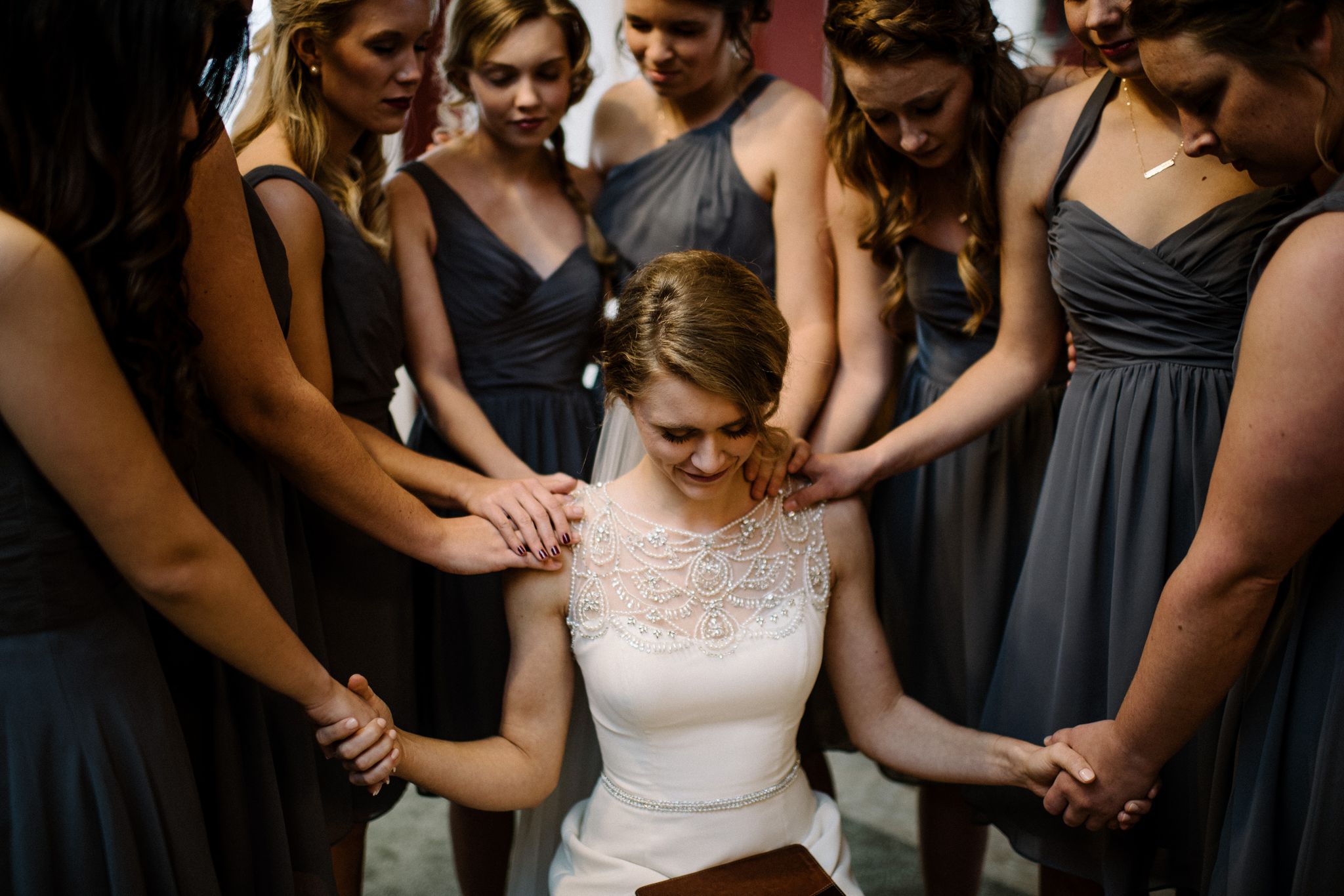 A closeup of the bride holding the hands of two of her bridesmaids. The rest of the bridesmaids have their hands on her shoulders. They are all praying together. 