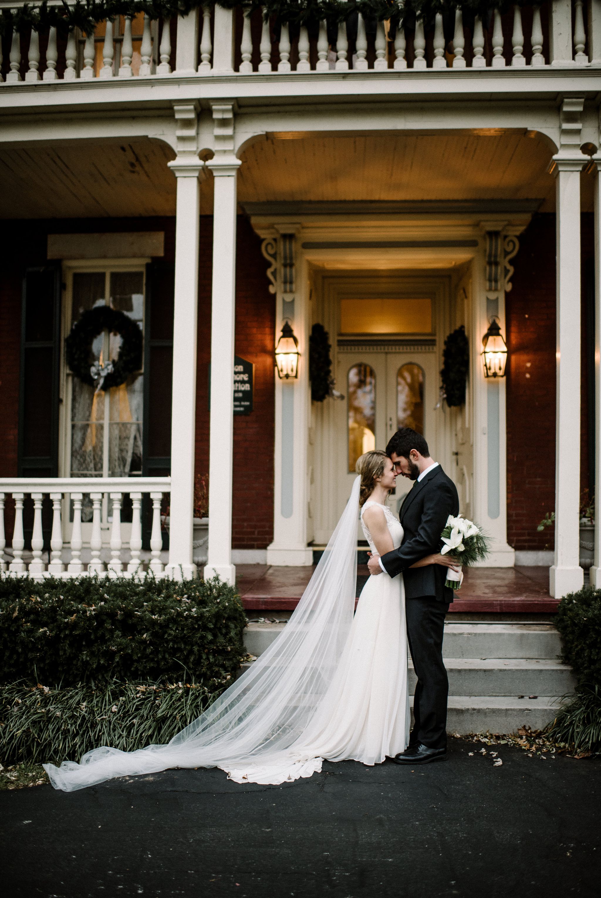 The bride and groom stand face to face near the front steps of the Larimore House. There are wreaths on the windows and front door. The bride is clutching her green and white bouquet. They are gazing into each others' eyes. 