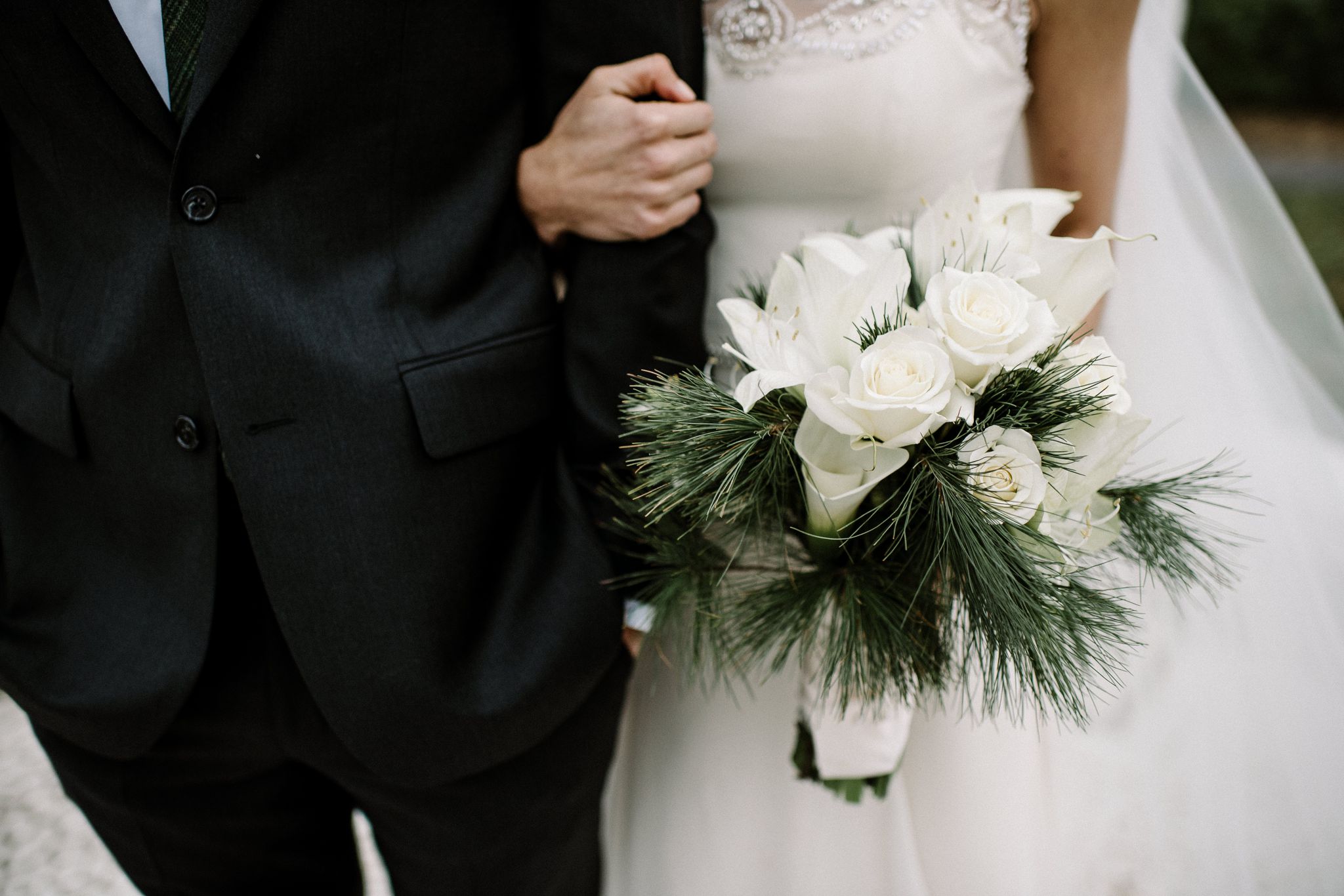 A closeup of the bride's bouquet of white roses and white lilies with greenery. Her arm is linked through the groom's arm. His hand is in his pocket.