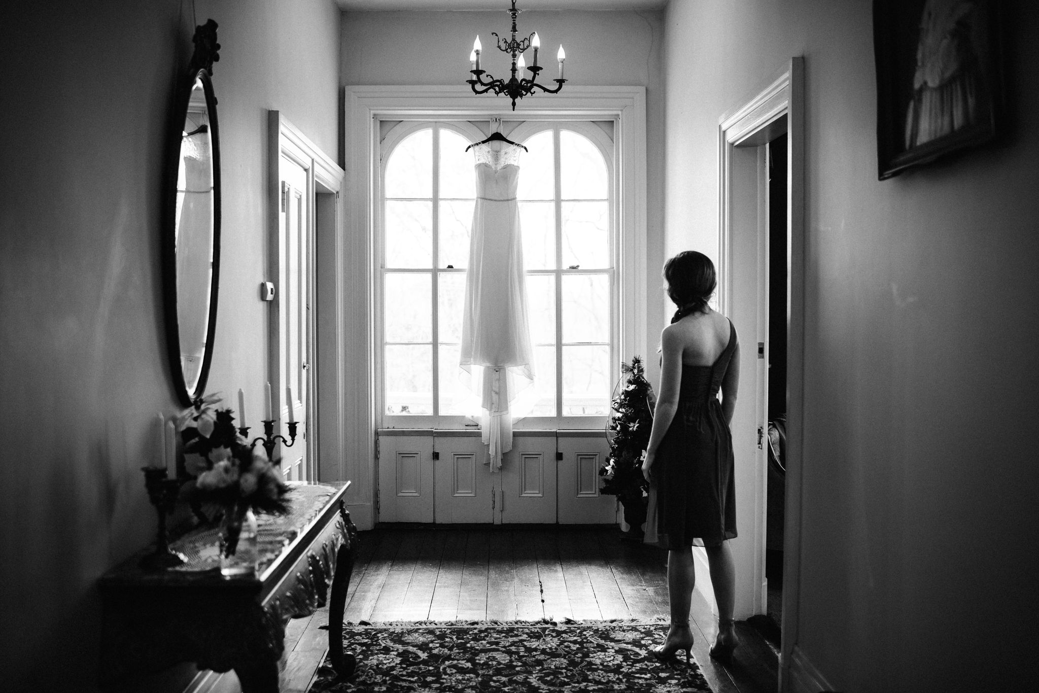The bride's sister is standing in the doorway, admiring the simple wedding gown hanging in the window at the end of the hall at the Larimore House. 