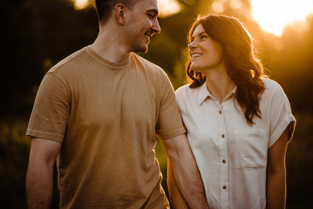 a couple walks towards the camera, smiling at each other during their golden hour engagement session. The woman is saying something to the man. 