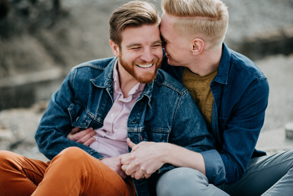 same-sex male couple sitting next to each other. they are smiling and their arms are around each other. the man on the right has his head nuzzled into the other mans forehead. they are happy.