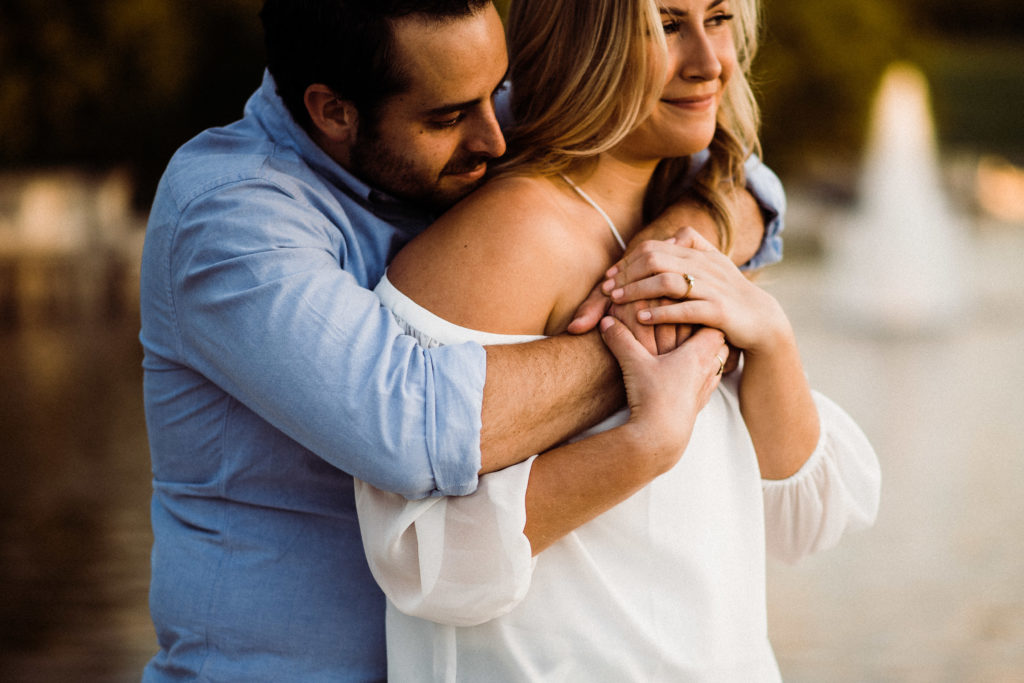 A man hugs his arms around his fiance from behind. He gently kisses her shoulder as she looks off into the distance during their golden hour engagement session.