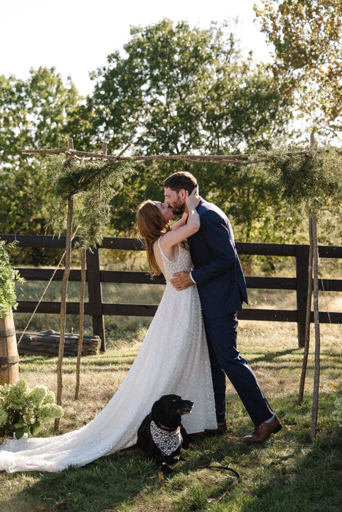 A couple dressed in a wedding gown and suit kiss under a rustic wooden arch outdoors. A dog wearing a decorative collar sits at their feet, and trees and greenery surround them in the background—perfect inspiration for elopement celebration ideas.