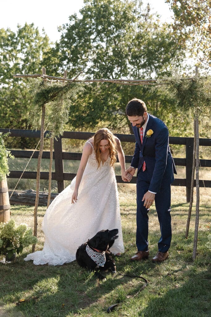 A bride and groom, holding hands, bend down to a black dog wearing a decorated bandana while standing under a wooden arch adorned with greenery. They are outside in a grassy area with trees and a fence—truly one of the most heartwarming elopement celebration ideas.