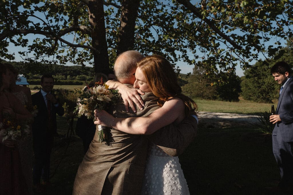 A bride in a white dress hugs an older man in a brown suit outdoors under a tree while holding a bouquet, capturing one of the many beautiful moments from their intimate elopement celebration.