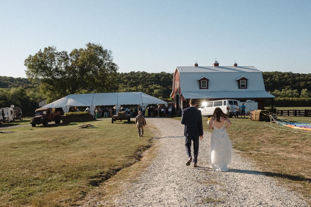 A couple walks towards a barn and a white tent where an elopement celebration is taking place, with greenery and trees in the background, on a sunny day.