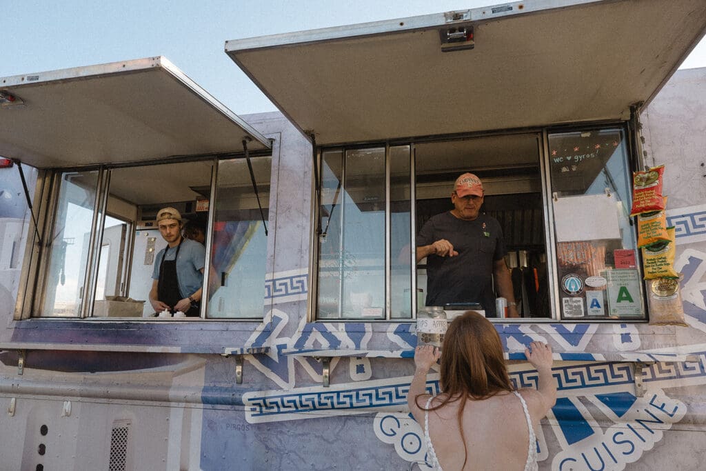 A person stands at the counter of a food truck, engaging with two employees inside. The food truck, adorned with elopement celebration ideas, boasts a vibrant graphic design and various items displayed on its exterior.