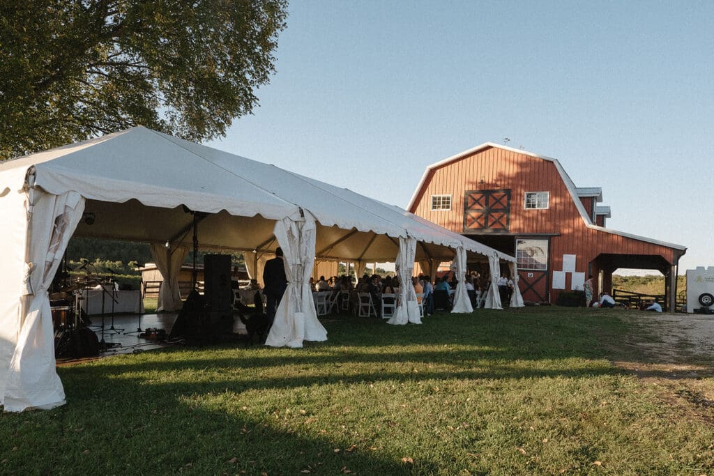 A white event tent is set up near a red barn with people gathered underneath, perfect for elopement celebration ideas. The scene is outdoors on a clear day with green grass in the foreground.