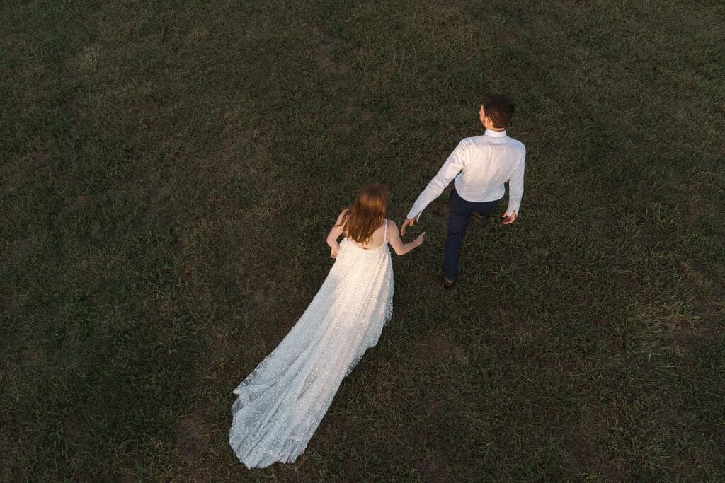 A couple, seen from above, walks hand in hand across a grassy field. The woman wears a long white dress and the man is dressed in a light-colored shirt and dark pants—an idyllic scene perfect for your elopement celebration ideas.