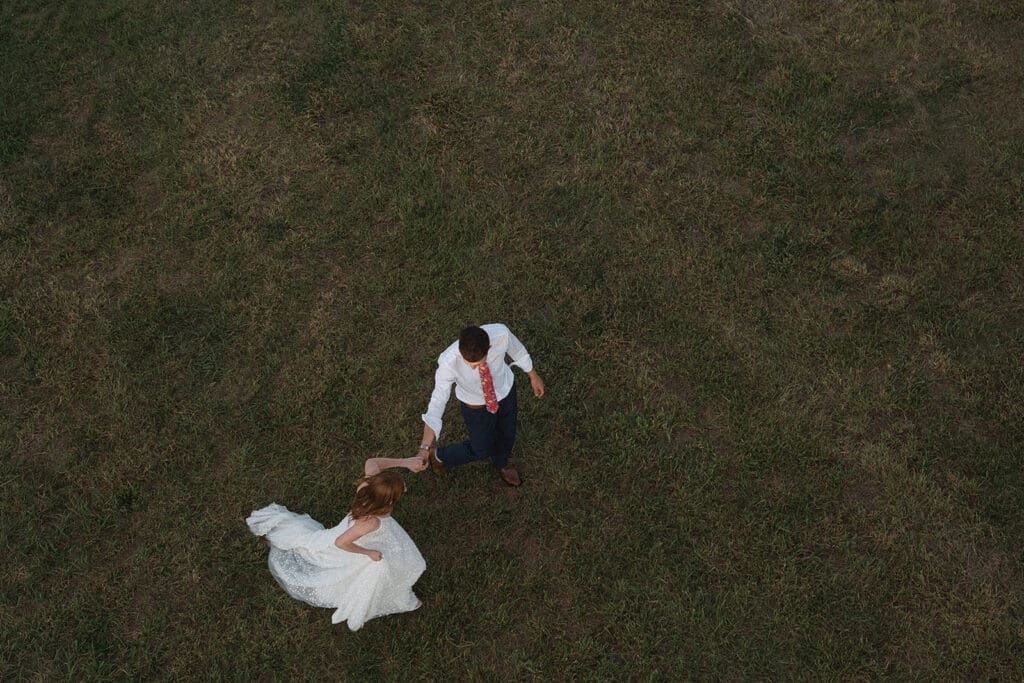 Aerial view of a bride in a white dress and a groom in a white shirt with a red tie dancing on a grassy field, perfect for those seeking unique elopement celebration ideas.