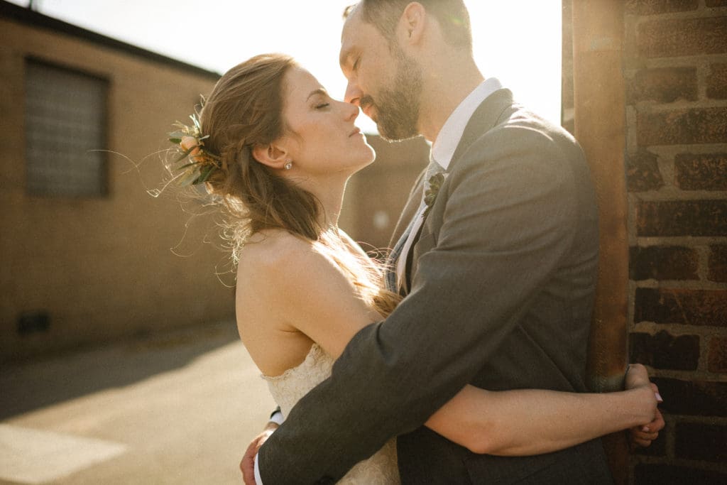 the groom is leaning agains the corner of a brick building. he is embracing the bride and they are almost kissing. the sun is shining just over his shoulder.