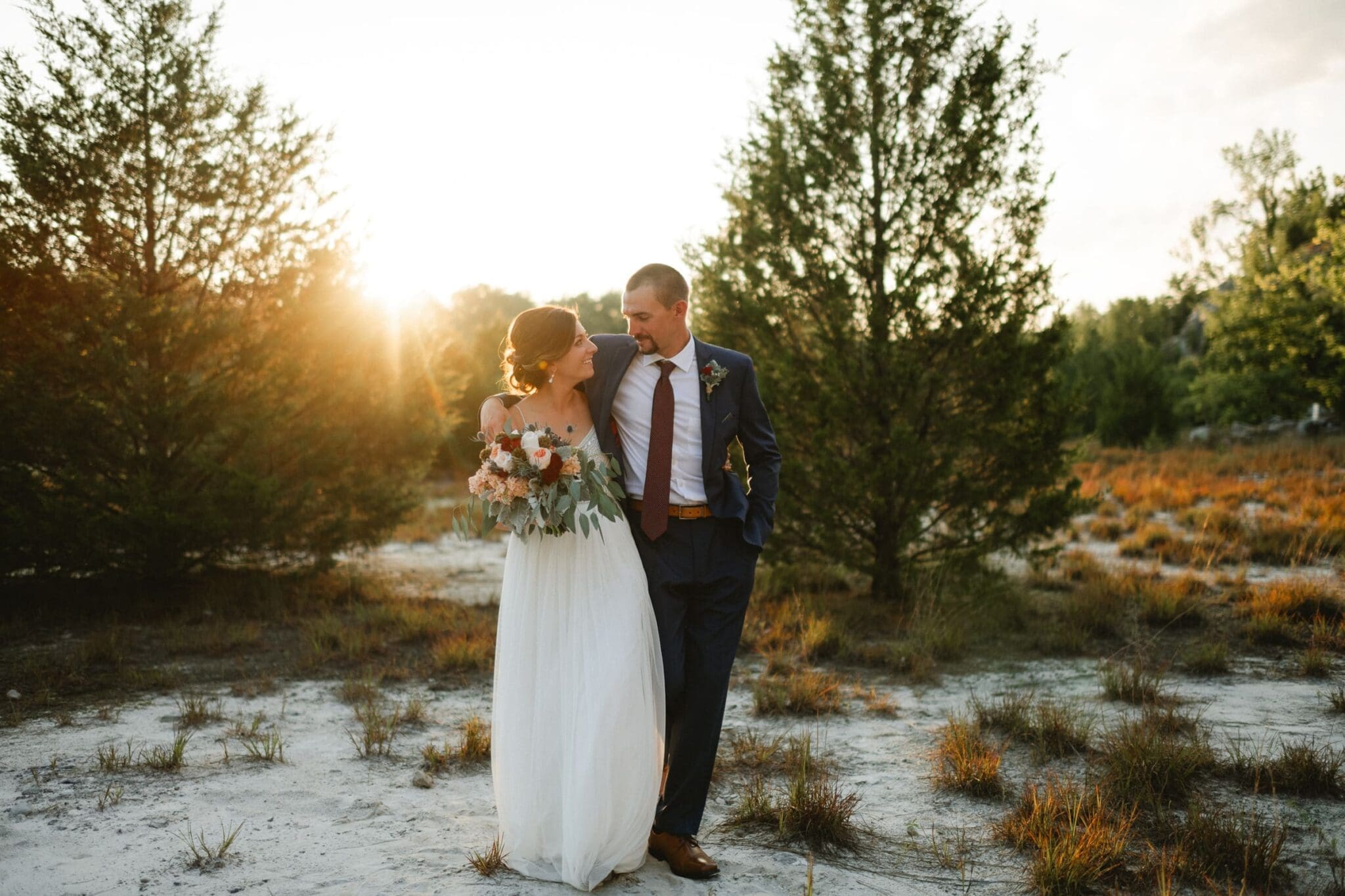 A bride and groom walk arm in arm in a natural, outdoor setting at sunset, with the groom in a suit and the bride in a white dress holding a bouquet—capturing the essence of elopement photography.