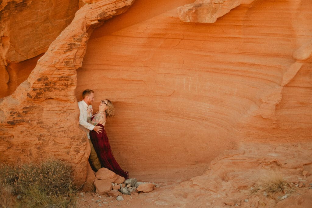 A couple stands in a rocky, orange desert landscape, with the man leaning against a rock formation and the woman leaning into him, ready to break the norms at your elopement.