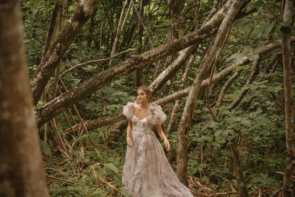 A woman in a floral dress walks through a dense forest with leaning trees and lush greenery—truly breaking the norms at your elopement.