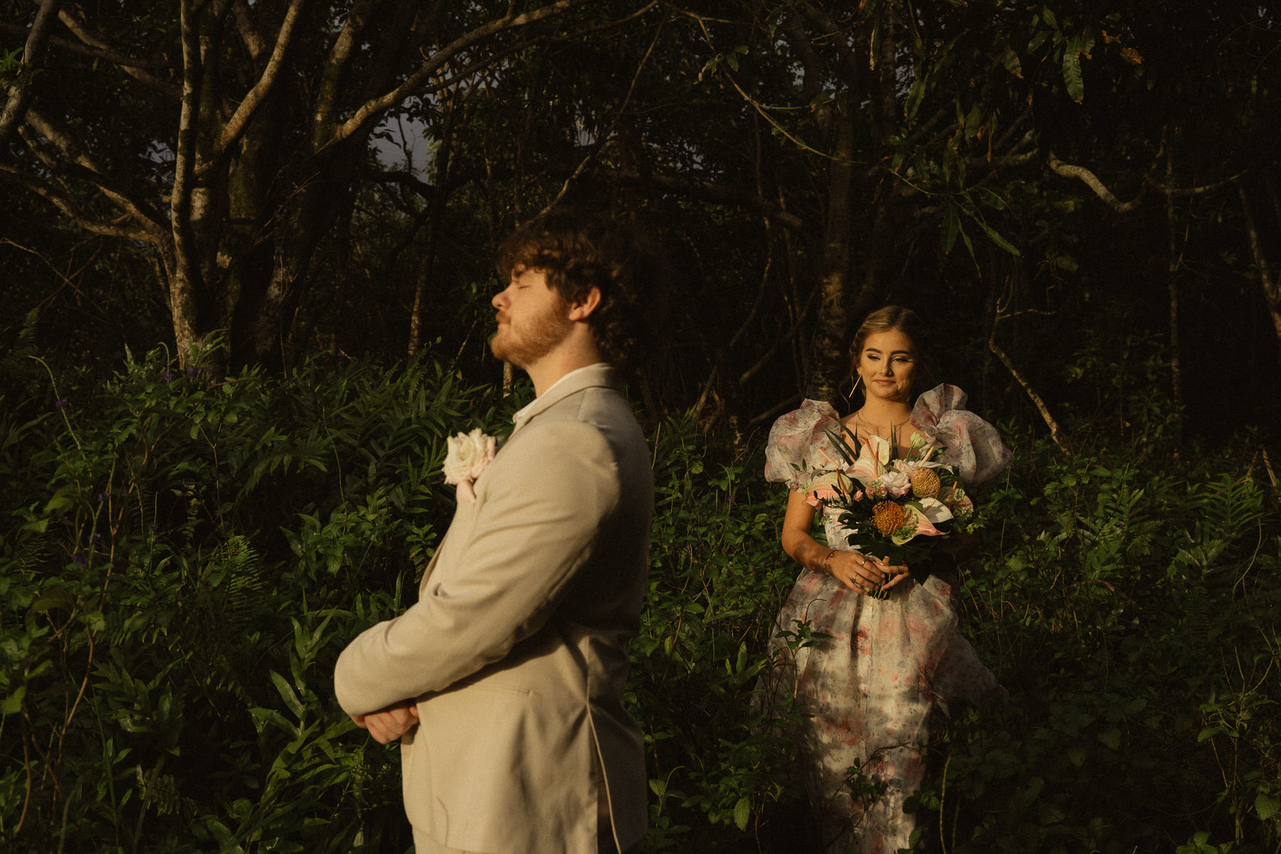 A man in a light beige suit stands with his arms crossed and eyes closed while a woman in a floral dress holding a bouquet stands behind him in a forested area, ready to break the norms at their elopement.