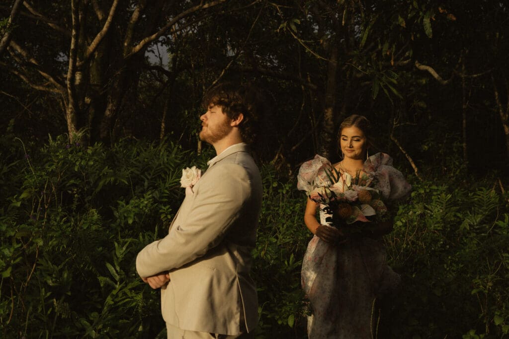 A man in a light-colored suit stands with his arms crossed in front of a woman in a floral dress holding a bouquet, both amidst the serene beauty of the forest. Together, they break the norms at their elopement, celebrating love in their unique way.