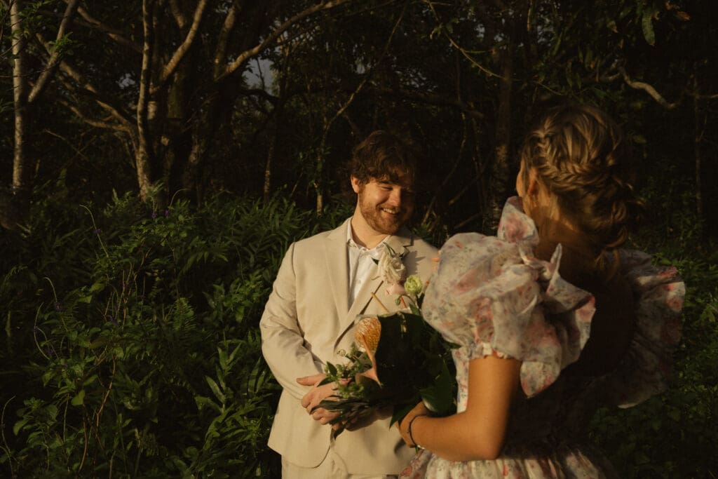 A man in a light beige suit and tie smiling at a woman with braided hair in a floral dress, holding a bouquet, standing in a forested area. They seem to break the norms at their elopement with their unconventional yet enchanting setting.