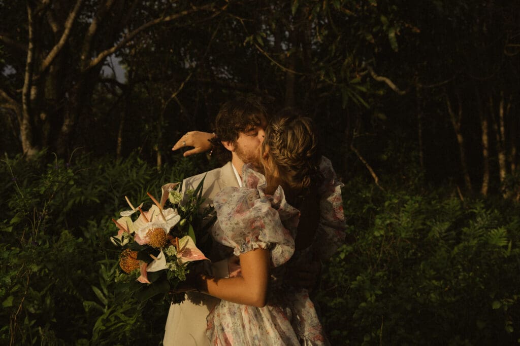 A couple in floral attire embraces and kisses outdoors, surrounded by dense greenery. One person is holding a bouquet, choosing to break the norms at their elopement.