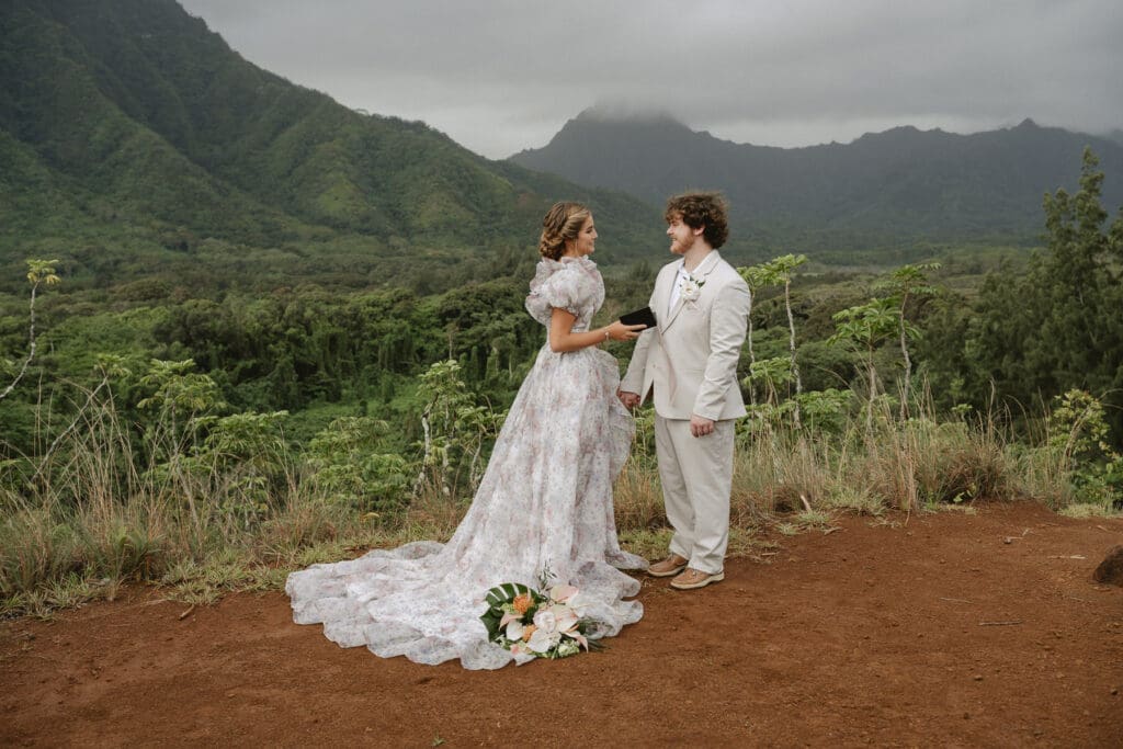 A couple, dressed in formal attire, stands on a scenic mountaintop with lush green hills and cloudy skies in the background. A bouquet of flowers lies on the ground, as they break the norms at your elopement for a more authentic celebration.