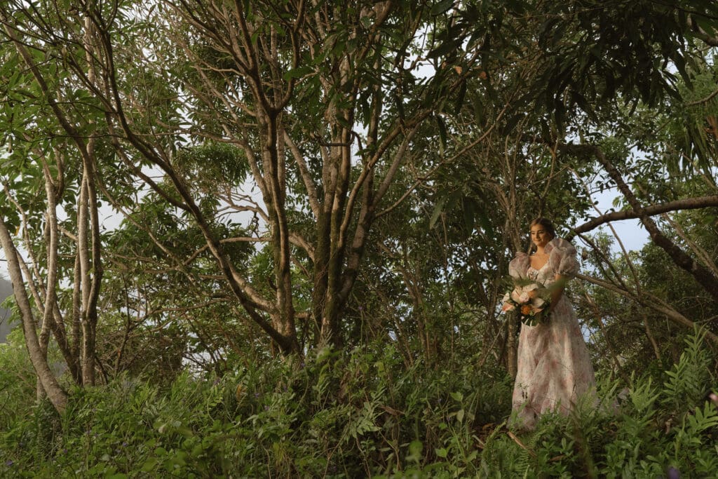 A person in a floral dress standing among tall trees and bushes, holding a bouquet of flowers, ready to break the norms at her elopement.