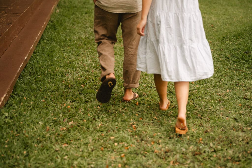 Two individuals walking away on a grassy surface; one is wearing brown pants and sandals, the other a white dress and sandals. They break the norms at your elopement.