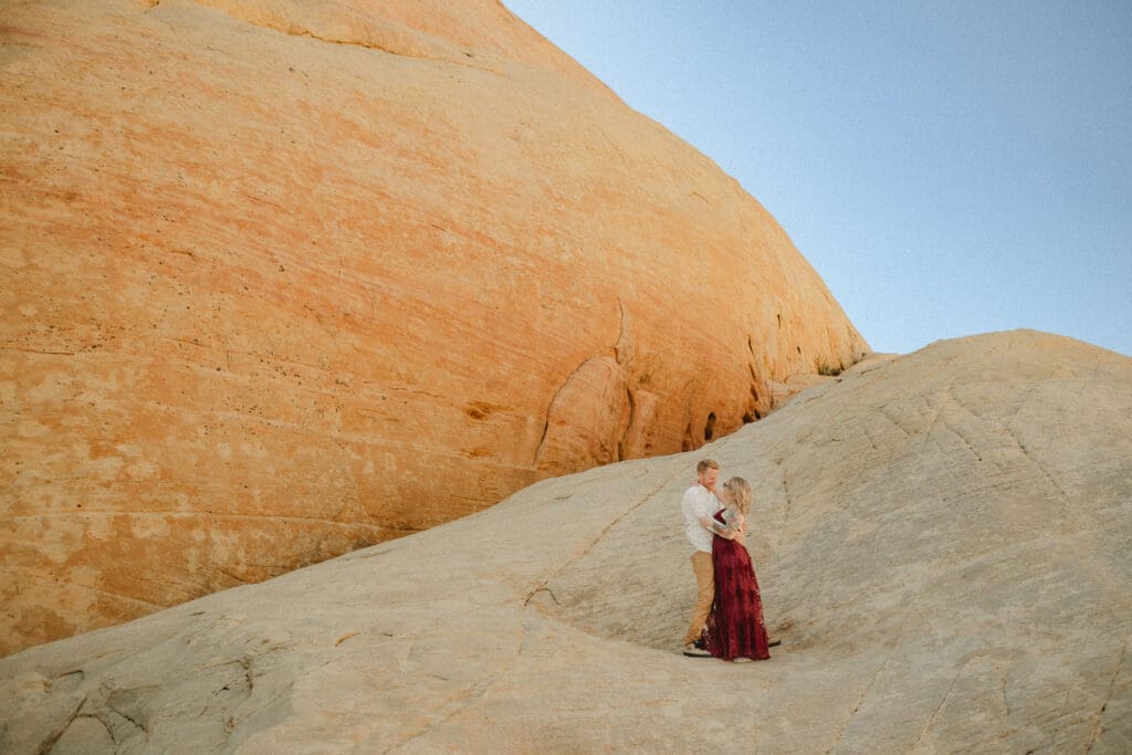 A couple stands on a rock formation in a desert landscape during daylight, with the woman wearing a red dress and the man in light-colored clothing, breaking the norms at your elopement.