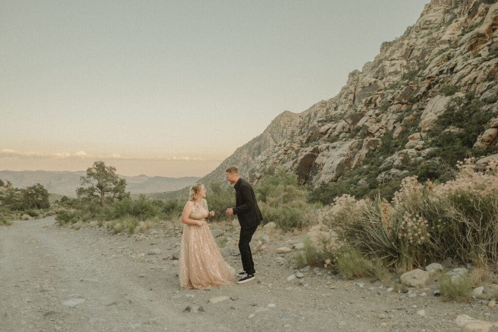 A couple, dressed formally, shares a moment on a rocky path in a mountainous landscape during sunset. The woman wears a pink gown while the man is in a dark suit. Break the norms at your elopement and embrace spontaneity with such breathtaking backdrops.