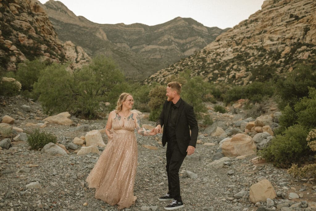 A man in a black suit and a woman in a light, sequined dress hold hands while walking in a rocky, mountainous landscape, breaking the norms at your elopement.