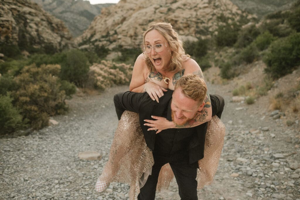 A person gives a piggyback ride to another on a rocky trail surrounded by hills and shrubs, both are smiling and laughing, breaking the norms at your elopement with pure joy and spontaneity.