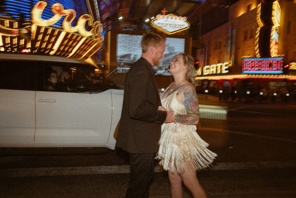 A couple stands on a city street at night, illuminated by surrounding neon signs. The woman wears a fringed dress and the man wears a dark suit. They are holding hands and smiling at each other, ready to break the norms at your elopement.