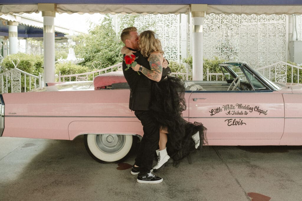 A couple embraces in front of a pink convertible with "Little White Wedding Chapel" written on the door. The woman, breaking the norms at your elopement, wears a black dress and white boots while holding a red bouquet.