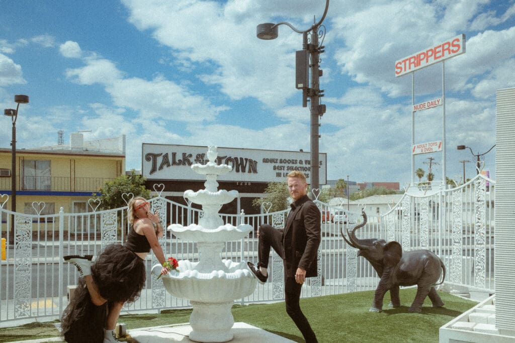 Two people in black outfits posing with a white fountain in an outdoor area decorated with an elephant statue and neon signs, ready to break the norms at your elopement.